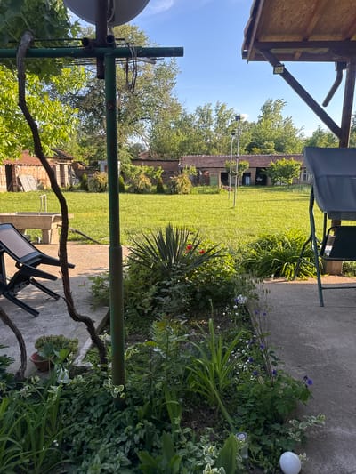 The image depicts a backyard scene in Lehchevo, Bulgaria. The setting appears to be a residential garden with a large grassy lawn extending towards various buildings in the background. The foreground features an outdoor sitting area with chairs and tables positioned on a concrete patio. Abundant vegetation, including flowering plants and shrubs, grows alongside the patio. The lighting indicates a sunny day, and the overall scene conveys a tranquil and natural environment typical of a village setting in Bulgaria.