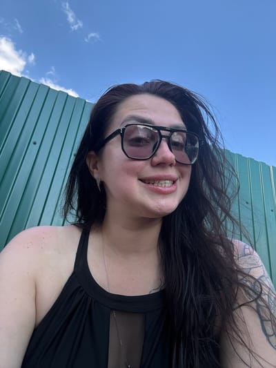 A medium shot, eye-level selfie of a woman with long, dark hair, wearing large, dark-rimmed sunglasses and a black top with a mesh insert. She is smiling broadly, showing her teeth. She has a winged earring in her left ear and a tattoo on her right arm. Behind her is a tall, green corrugated metal fence, and above that, a clear blue sky with a few scattered white clouds. The scene appears to be outdoors, possibly in a park or a backyard. No specific location in the city of Узюково, Russia can be identified from the image.