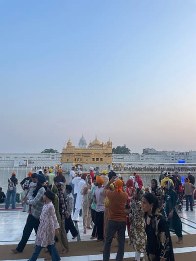 The image depicts a crowd of people gathered at the Golden Temple, also known as Harmandir Sahib, in Amritsar, India. The iconic golden structure of the temple is visible in the background, surrounded by a reflective pool of water. The sky is a clear, pale blue, suggesting it might be early morning or late afternoon. People are seen walking, standing, and interacting, many wearing traditional Sikh attire, including turbans. The scene appears to be bustling with activity, as visitors engage with the sacred site. The foreground shows a paved walkway where individuals are moving in various directions.