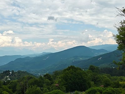 This image depicts a wide, elevated view of a verdant mountain landscape under a cloudy sky. The foreground is dominated by lush green trees and foliage, with some of the trees extending into the upper right corner of the frame. Beyond the immediate trees, the terrain slopes downwards, revealing rolling hills covered in dense vegetation. In the mid-ground, scattered houses and small settlements are visible nestled amongst the hills, suggesting a rural or semi-rural environment. Further in the distance, layers of mountains recede into a hazy blue, with snow-capped peaks visible on the horizon. The sky is filled with a mix of white and gray clouds, with patches of blue sky peeking through. The overall impression is one of a vast, natural landscape with signs of human habitation. The location is identified as Ukeshi, Georgia.