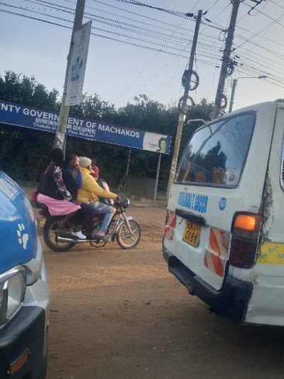 The scene depicts an unpaved road in Mua ward, Kenya. A silver-colored motorcycle, bearing three individuals, occupies the mid-ground. The driver wears a yellow long-sleeved top and blue jeans. The middle passenger wears a dark patterned top and a white head covering. The rear passenger wears a dark top and a pink skirt. To the right of the motorcycle, the rear of a white commercial van is visible, displaying license plate "KAY 018K", orange and white reflective hazard stripes, and an illuminated orange right signal light. Text "ASSISTANT COACH" is partially visible on its upper rear. On the left, a partial view of a blue/grey vehicle with a white paw print decal is present in the foreground. In the background, a large blue banner identifying the "COUNTY GOVERNMENT OF MACHAKOS" and "DEPARTMENT OF HOME DEVELOPMENT" hangs against a backdrop of dense green foliage. Multiple utility poles with overhead wires are present. The sky is light blue.