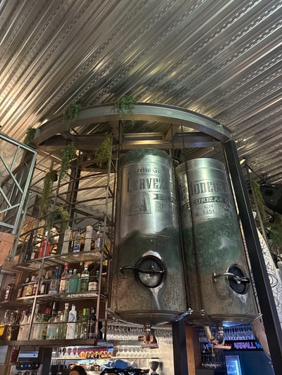 The image depicts the interior of a bar or restaurant in Port D'alcúdia, Spain. The scene is dominated by a large, industrial-style metal structure that holds two substantial cylindrical tanks, likely for storing beverages. These tanks are tarnished and show signs of age, with some green patination, and feature circular access hatches with handles. Hanging plants cascade from the metal framework above, adding a touch of greenery to the otherwise metallic and utilitarian design.
To the left of the tanks is a multi-tiered shelving unit laden with numerous bottles of alcohol, including various spirits and liqueurs. The shelves are made of metal, and the bottles are arranged in a somewhat organized but dense fashion. Below the shelving unit, part of a bar counter is visible, with a coffee machine and other bar equipment. In the background, rows of glassware, likely wine or cocktail glasses, are hung upside down from racks.
The ceiling is constructed from corrugated metal, reflecting the lighting in the space. The overall aesthetic suggests a modern industrial or rustic theme, possibly a craft brewery or a bar with a focus on unique beverage presentation. There are no identifiable people clearly visible or engaged in any action in the frame. The lighting appears to be ambient, casting some shadows and highlighting the textures of the metal and glass.