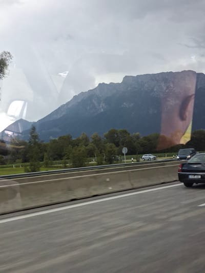 This image captures a roadside view of a mountainous landscape on a cloudy day, likely taken from a vehicle traveling on a highway. The foreground features a grey concrete highway barrier and the asphalt road, with white lane markings visible. In the middle ground, several cars are on the highway, including a black sedan in the right lane and a silver SUV in the left lane. Beyond the highway, a dense line of green trees separates the road from a dark, imposing mountain range that dominates the background. The sky is overcast, with grey clouds obscuring the sun. The image appears to be taken in Oberaudorf, Germany, a region known for its scenic mountain vistas. A reflection of a person's face is visible on the glass surface through which the photo was taken.