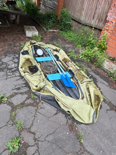 In Kalitino, Russia, an inflated, olive-green inflatable boat rests on a cracked asphalt surface. The boat is partially covered with dirt and debris. Inside the boat, a circular, silver pressure gauge is visible, along with two blue oars and a coiled rope. The boat is situated outdoors, with a wooden fence and brick wall in the background, overgrown with various green plants and weeds.