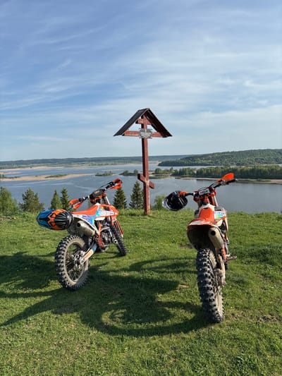 Two orange dirt bikes are parked on a grassy hill overlooking a body of water in Elkino, Russia. The foreground features a wooden directional sign. Both bikes have helmets resting on their handlebars. The sky is partly cloudy.