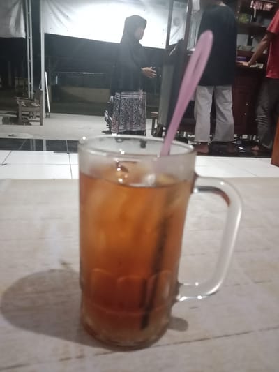 A glass of iced tea with a pink straw is in the foreground, placed on a light-colored, textured surface. The glass has a handle and is filled with a dark amber liquid and ice cubes. In the background, a woman wearing a black hijab and a patterned skirt stands near a wooden cart. To her right, a person in a black t-shirt and light-colored pants, and another person wearing a red t-shirt and dark pants are visible. They appear to be in an outdoor area with a white canopy overhead. The ground is paved with tiles, and there is a wooden chair to the left. The overall lighting suggests it is nighttime or dusk. The location is identified as Cangkring, Indonesia.