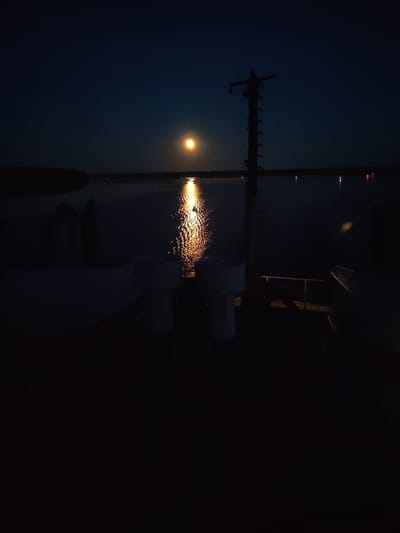 The image shows a night scene in Irdomatka, Russia. A body of water reflects moonlight, creating a bright streak across the surface. On the water's edge, faint lights are visible on the opposite shore. A dark, silhouetted structure resembling a mast stands to the right of the moonlight reflection. The foreground is dark, but shows the deck of a boat.