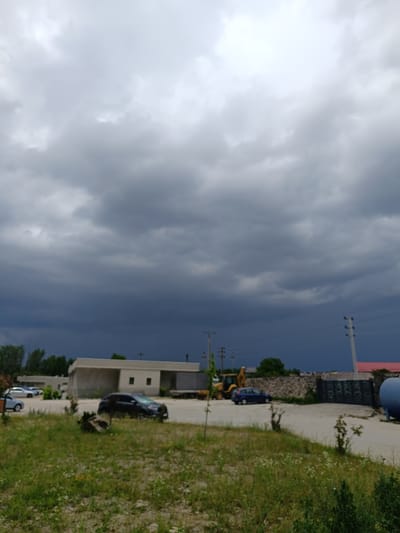 The image depicts an outdoor scene under a dramatic, overcast sky with dark storm clouds gathering. The setting appears to be a semi-industrial or construction area, possibly on the outskirts of Târgu Jiu, Romania.

In the foreground, there is a patch of unkempt grass with scattered wildflowers, suggesting a natural, undeveloped area bordering the built environment.

Further back, a light-colored, low-rise building with a flat roof is visible. In front of the building, several cars are parked on a paved surface. A dark-colored SUV is prominently featured in the mid-ground, while other vehicles, including a lighter-colored car and potentially others, are partially visible.

An excavator, a yellow construction vehicle, is parked in the middle ground, alongside a blue sedan. A dark metal gate and a large blue cylindrical tank are also present, hinting at industrial or storage activities. Power lines and utility poles are visible in the distance, further emphasizing the semi-industrial nature of the location.

The overall mood is somber and anticipatory, largely due to the menacing storm clouds dominating the sky. The time of day is ambiguous but could be late afternoon or early evening, given the intensity of the clouds. There are no people visible in the image. No visible text is present.
