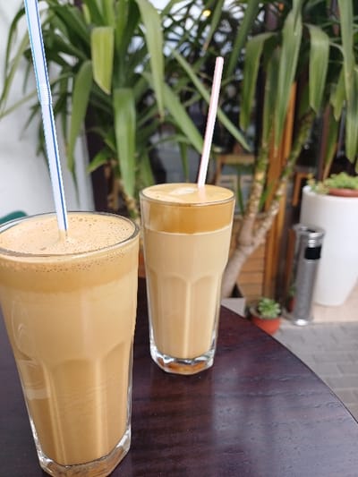 Two tall, clear glasses containing beverages are centrally positioned on a dark brown, rounded table in an outdoor cafe setting in Gevgelija, North Macedonia. The glass on the left holds a frothy, light brown beverage with a blue and white striped plastic straw inserted. The glass on the right, positioned slightly behind, contains a layered beverage with a lighter brown liquid base and a darker foam top, with a pink plastic straw. The background features several large green plants with long, pointed leaves. To the far right, a silver cylindrical object, likely a standing ashtray or patio heater, is visible next to a white planter holding a small green plant. The ground beyond the table is paved with light-colored square tiles. No individuals are present in the scene.