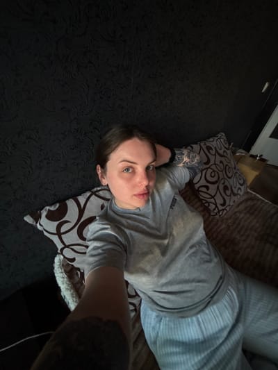 A person with short brown hair and fair skin lies on a bed in a room. The person is wearing a grey t-shirt and striped pajama pants. The bed has brown bedding and two pillows with brown and white patterns. The floor is a dark color with a subtle pattern. A portion of a dark-colored piece of furniture is visible in the upper left. The city of Chornomorskoe, Ukraine, is not apparent in the scene.