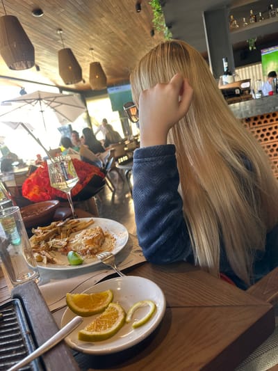 A blonde-haired person is seated at a wooden table, an arm resting next to a white plate containing a few orange slices. Another plate holds remnants of a meal, accompanied by a fork. A partially visible bottle and glass are also on the table. In the background, other individuals are seated under umbrellas, and a television screen displays an image. The setting appears to be an outdoor patio or restaurant.