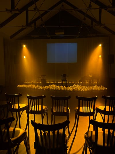 The image depicts a dimly lit performance space, possibly a small theatre or auditorium, bathed in a warm, golden light emanating from spotlights. The foreground is occupied by rows of dark wooden chairs with slatted backs, casting long shadows on the floor. In the midground, a vast sea of unlit tea lights covers the floor, creating a textured, candlelit landscape. Beyond the tea lights, on a raised platform, stand several objects: a grand piano with what appear to be a few lit tea lights on its surface, a music stand with a book, and a single, empty chair. Behind these elements, a large projection screen displays a muted blue rectangular image. The ceiling structure is visible, with exposed beams and track lighting. Curtains hang on either side of the projection screen, and a speaker is positioned on the right. The overall atmosphere is serene and contemplative, suggesting a staged event or memorial. The location in Ilyanskiy selskiy Sovet, Belarus, is not discernible from the visual information present in the image.