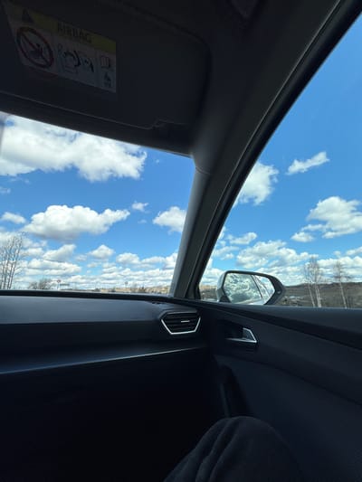 The image, originally rotated 90 degrees counter-clockwise, displays a vibrant view from inside a car looking outwards on a bright, clear day in Печмень, Russia. The vast blue sky, adorned with numerous fluffy white cumulus clouds, dominates the upper and central portions of the frame.
The car's interior is visible along the bottom and sides. On the left, the dark fabric of the vehicle's headliner and the edge of a sun visor are present. An "AIRBAG" warning label, along with safety pictograms illustrating proper child and adult seating, and a symbol prohibiting rear-facing child seats in the front, is clearly visible on the sun visor.
To the right, parts of the car's dashboard and door panel are seen, including a modern air vent with a metallic-looking trim. A portion of a dark-colored garment or seat suggests the presence of a passenger. The car's exterior side mirror is visible in the upper right quadrant, reflecting the cloudy sky and the distant landscape.
The landscape visible beyond the car appears to be a mix of bare trees, characteristic of early spring or late autumn, and some distant features, suggesting a rural or suburban setting. The bright sunlight indicates daytime. The image captures a serene moment of travel or contemplation while looking out at the open sky.