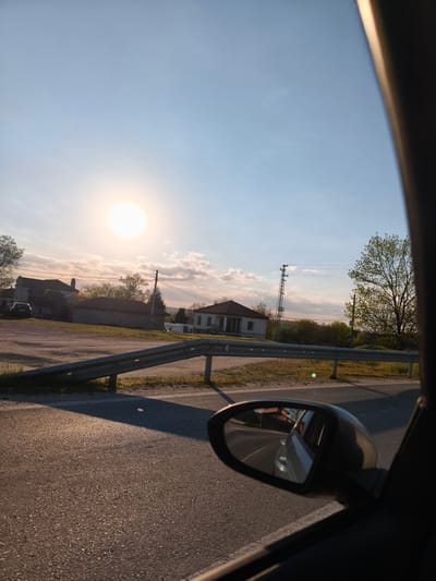 The image shows a view from inside a car, likely on a road in or near the village of Zdravets, Bulgaria. The bright sun dominates the sky. A road stretches out in front, with a metal guardrail along the side. Beyond the guardrail, there's a grassy area and several houses in the distance, some with dark roofs. A power line pole is also visible. The side mirror of the car is in view, reflecting the road and another car. A portion of the car's interior frame is visible on the right side of the image.