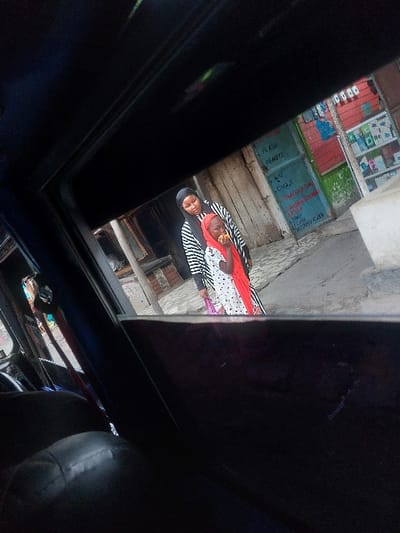 The image depicts an urban street scene in Mwandege, Tanzania, viewed from inside a vehicle. Two individuals stand on a light-colored ground surface in the mid-ground. An adult female, wearing a black head covering and a black and white striped long-sleeved garment, stands slightly behind and to the right of a child. The child, likely a girl, wears a red head covering and a white garment with dark spots, and is actively eating a yellow fruit. The adult female holds a purple plastic bag in her left hand. To the right of the individuals, a building façade features two doorways; the left door is light-colored and plain, while the right door is green and bears white hand-painted text advertising items or services such as "FLASH REMOTE," "WIRE CHAJI," and "TOFAUTI SUMI." Above and to the right of the green door, a shop display area contains various small packaged goods on shelves with red backing. Indistinct building structures are visible in the background to the left.