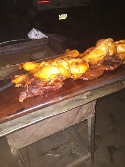 Grilled meat, chicken, and other foods sit on a wooden cutting board. The board is on top of a metal table, which is covered with a worn, tan fabric. The table is in a dimly lit outdoor setting. In the background, there is a car with its taillights on and a license plate that is visible. There is also crumpled paper, probably containing leftovers.