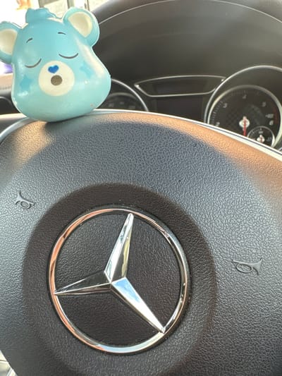 The image displays a light blue, bear-shaped air freshener or figurine positioned on the dashboard of a Mercedes-Benz vehicle, assumed to be located in Easingwold, United Kingdom. The bear has closed eyes, a small blue heart on its chest, and cream-colored inner ears. Directly below it, the black, textured steering wheel is prominent, featuring a central silver Mercedes-Benz three-pointed star logo. Two small horn symbols are visible on the steering wheel, one to the left and one to the right of the central hub. In the background, portions of the vehicle's instrument cluster, including a speedometer or tachometer dial, are visible. The scene is illuminated by natural light entering from an unseen source, creating highlights on the bear figure and the metallic Mercedes-Benz emblem. No individuals are present, and no actions or interactions are depicted.