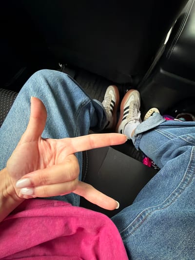 In a dimly lit vehicle interior, the image captures a close-up view of a person's hands and legs, suggesting they are seated. The person is wearing blue denim jeans and white sneakers with gum soles and black stripes. A bright pink garment, possibly a shirt or jacket, is visible in the foreground. The hand positioned in the center of the frame makes a "rock on" hand gesture, with the thumb and index finger extended and the other fingers curled. The background consists of dark upholstery, likely car seats. The scene's low lighting and the specific details of the clothing and hand gesture suggest an informal, candid moment. The image does not contain any discernible elements that would place it in Kimovsk, Russia.