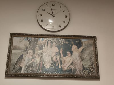 The image shows a framed painting hanging on a white wall, with a clock mounted above it. The painting depicts a religious scene featuring Mary holding the infant Jesus, surrounded by several angels. Mary is in the center, wearing a blue and white robe, and cradling Jesus, who is naked. To the left of Mary, an angel with large wings is looking towards her. To her right, another angel offers a flower to Jesus. A smaller angel stands to Mary's left, and another to her right. The painting is framed in an ornate gold-colored frame. The clock above the painting is round with Roman numerals and black hands. The clock indicates the time is approximately 10:55. The location is identified as the city of Ocnița, Moldova.
