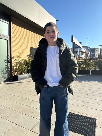 A male, appearing to be a juvenile, stands centrally in an outdoor urban setting in Lipomo, Italy, under a clear blue sky. He is wearing a dark olive green hooded puffer jacket over a white t-shirt featuring a small red emblem on the left chest pocket, and blue denim jeans with his hands positioned in the front pockets. The subject's gaze is directed forward towards the camera. He stands on light-colored rectangular paving stones. To his left, a modern building with light brown brickwork and a dark-framed window is visible, casting a distinct shadow. To the subject's right and in the background, green shrubbery in rectangular planters is present, along with additional urban buildings and part of a large road or commercial sign. A black metal gridded drain cover is located on the pavement to the subject's lower right.