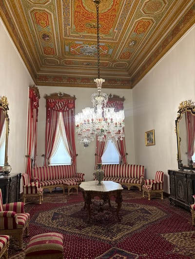 The image captures an opulent interior of what appears to be a drawing-room in Üsküdar, Turkey. The scene is dominated by rich, detailed decor. A grand, ornate chandelier made of crystal hangs from a meticulously painted ceiling featuring intricate gold patterns and colorful motifs. The walls are adorned with red and white striped curtains, some drawn back to reveal bright daylight, suggesting the time of day is likely daytime.
The room is furnished with plush seating arranged around a table. The furniture is upholstered in red and white stripes, with gilded frames on some of the chairs. A large, intricate rug with a red base and patterned designs in beige, blue, and black covers the floor. A dark, carved wooden cabinet stands against one wall, topped with decorative elements. A gilded mirror with ornate scrollwork is also visible, reflecting the room. A framed picture hangs on the wall, though its details are indistinct.
The overall impression is one of grandeur and historical richness, suggesting a well-preserved or restored traditional Turkish interior. There is no visible text in the image. The setting evokes a sense of timeless elegance and tranquility.