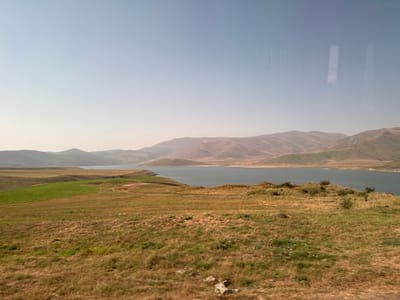 The image displays an expansive outdoor landscape, featuring a large body of water, identified as being in the vicinity of Tsghuk, Armenia. The upper two-fifths of the frame consist of a light blue sky with a distinct atmospheric haze. The foreground is primarily composed of dry, golden-brown grasses and sparse vegetation, with patches of greener growth towards the left and scattered small rocks visible at the bottom. The midground is dominated by the dark blue-green water body, exhibiting an irregular shoreline with visible peninsulas extending into it. In the background, a series of undulating hills and mountains, characterized by earthy tones of brown, green, and grey, recede into the distance, partially obscured by the pervasive atmospheric haze. No individuals, animals, or prominent man-made structures are present within the depicted natural environment.