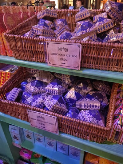 The image shows two wicker baskets filled with blue and gold, pyramid-shaped boxes labeled as "Harry Potter MILK CHOCOLATE FROG" and "Harry Potter WHITE CHOCOLATE FROG" with price tags of £9.95. The baskets are located on shelves in a shop. In the top basket are boxes of milk chocolate frogs. The lower basket contains boxes of white chocolate frogs and small boxes of toad candies. The location appears to be a Harry Potter-themed shop, likely in the city of Three Rivers, United Kingdom. There are people in the background, but their faces are not distinct.