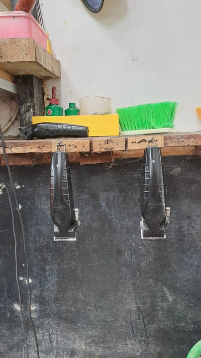 The image depicts a close-up, eye-level shot of a rustic shelf in what appears to be a barbershop. The shelf is constructed from weathered wooden planks, precariously attached to a white wall. A thick layer of dust and cobwebs covers the surfaces.
On the shelf, several items are visible: a yellow block, two small green bottles, one with a red cap and a white circular label bearing a red 'S', and another with a green cap. To the right of the bottles is a roll of white tape. A bright green bristled brush is positioned to the right of the tape. In front of the yellow block and bottles lies a black electric hair clipper.
Hanging from hooks on the underside of the shelf are two identical black electric hair clippers. These clippers are suspended by their handles, with their cutting heads facing downwards. The wall behind the shelf is painted white, with visible signs of wear and tear, including patches of missing paint and dust accumulation. The lower portion of the image shows a dark, textured surface, possibly a wall or backdrop, also showing signs of dust and grime. Black electrical cords are visible hanging down the left side of the frame.
Based on the context provided, this scene is located in the city of Kruengjuli Timu, Indonesia. The overall impression is one of a working, perhaps slightly unkempt, professional environment.