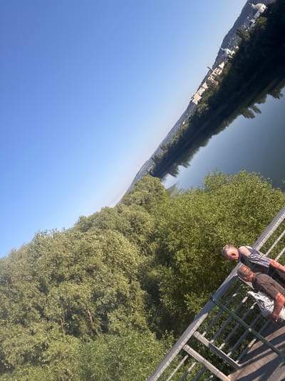 The image shows a view from a bridge overlooking the Dniester River in Otaci, Moldova. The river is calm and reflects the clear blue sky and the tree-lined banks. On the far bank, a town with white-domed buildings and some industrial structures is visible, nestled on a hillside covered with trees. Two men are standing on the bridge, looking out at the scenery. The man in the foreground is wearing a plaid shirt and the man behind him is wearing a dark shirt. Both appear to be older. The bridge has a metal railing and a mesh barrier. Lush green trees and foliage dominate the closer bank and the foreground of the image. The scene is bathed in bright sunlight.