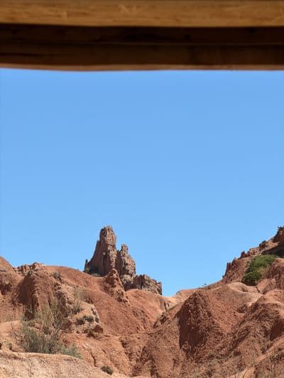 The image shows a view from Tosor, Kyrgyzstan, looking upwards at a reddish-brown, rocky cliff face against a clear blue sky. The cliff is textured and has some sparse vegetation growing on it. On the left side of the frame is a blurred wooden structure, possibly part of a building or frame. The photo is taken from a low angle, emphasizing the height and ruggedness of the terrain.
