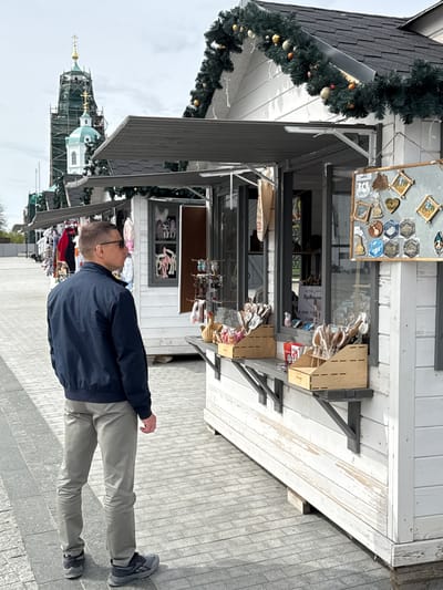 A man stands facing a kiosk on a paved street in Arzamas, Russia. He wears glasses, a dark jacket, and light-colored pants. The kiosk displays items for sale, including what appear to be crafts and souvenirs, arranged on shelves and displays. The kiosk is adorned with Christmas decorations. In the background, a building with a spire and scaffolding can be seen.