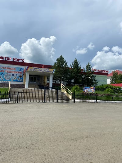 The image shows the exterior of a building labeled "ДОМ МОЛОДЕЖИ" (House of Youth) in Askarovo, Russia. The building has a red roof and is adorned with signs in Cyrillic script. To the left, a sign reads "СТАНЦИЯ ЮНЫХ ТЕХНИКОВ" (Station of Young Technicians). In front of the building, there is a black metal fence, a paved area, and some greenery. The sky is partly cloudy. There are no people visible in the image.