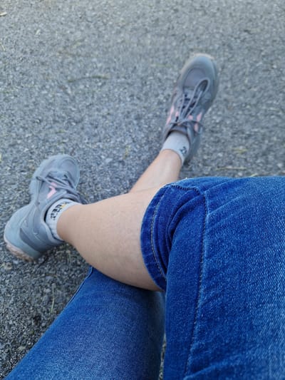 The image is a close-up shot of a person's legs, crossed at the knee, with both feet wearing gray athletic shoes with pink accents. The person is wearing blue denim shorts, and white socks that have a black and yellow logo on them. The background is a gravelly surface, likely an outdoor pathway. The lighting suggests it is daytime. The image was taken in Bahovitsa, Bulgaria.