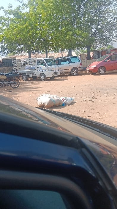 This daytime image, taken from within a dark blue vehicle, depicts an unpaved, dusty ground area with parked vehicles and vegetation in Langtang, Nigeria. In the immediate midground, two white or light-colored sacks are abandoned on the reddish-brown dirt surface. A black motorcycle with silver accents is visible on the left, partially obscured by the foreground vehicle. Beyond this, a white compact pickup truck with a caged cargo bed and blue decals is parked facing left, next to a light-colored minivan. Further to the right, a red compact minivan is parked. The background features several mature trees with dense green foliage under a clear, light blue sky. Behind the trees and vehicles, indistinct low-rise structures with corrugated roofs are visible. No individuals are present in the scene.