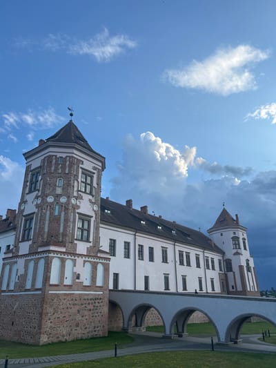 The image depicts Mir Castle Complex, a UNESCO World Heritage site located in the agrotown of Mir in the Karelichy District of Grodno Region, Belarus. The castle, with its distinctive towers and white walls, is seen under a dramatic sky filled with white and dark clouds. A bridge with arched supports connects different parts of the castle. Green grass and paved pathways surround the structure, suggesting an outdoor, well-maintained park or grounds. The architecture is a blend of Gothic, Renaissance, and Baroque styles. The sky suggests either early morning or late evening light, with a strong contrast between the brightly lit clouds and darker storm clouds.