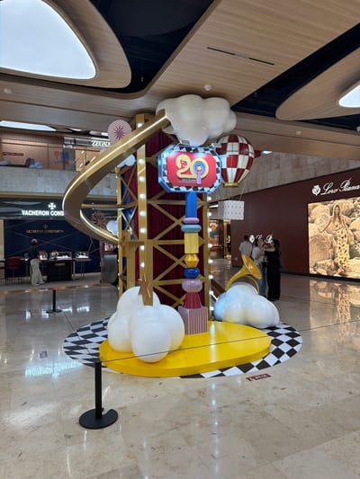 A festive, colorful display is set up in a well-lit, modern indoor shopping mall. The display features a central structure with a golden curved archway, adorned with lights and red velvet curtains. Several large, puffy white cloud-like objects are integrated into the display. Suspended above are two hot air balloons, one red and white checkered, and another with a geometric pattern. A prominent sign displays the number "20" in large, vibrant colors, indicating an anniversary celebration, with text "DEJI PLAZA 20TH ANNIVERSARY" below. The display sits on a checkered black and white circular platform. To the right of the display, a large digital billboard showcases a model in a patterned dress standing amongst rocks, with the brand "Loro Piana" displayed above. In the background, several people are visible. To the left of the display, the "VACHERON CONSTANTIN" boutique is identifiable, with display cases visible inside and a person browsing. The flooring is a light-colored marble with a reflective surface. The ceiling features modern, curved architectural elements with integrated lighting. The overall scene suggests a lively and celebratory atmosphere within a high-end retail environment, likely located in Xuanwu District, China, given the context of the Deji Plaza anniversary.