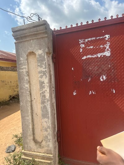 The image shows a section of a red diamond plate metal gate with some torn posters on it, attached to a grey concrete pillar. The pillar has a decorative recessed section and is weathered. Behind the pillar, a yellow wall is visible with a red-roofed building. The ground is sandy with some green vegetation and a black plastic bag. The sky is partly cloudy. A person's hand is visible in the bottom right corner, holding a light-colored rectangular object. This scene is located in Ogbomosho, Nigeria.