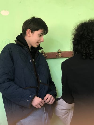 The image captures two individuals in an indoor setting with a light green wall as the background. While the location is specified as Ortabatumi, Georgia, no specific city landmarks are visible to confirm this within the image itself.
On the left, a young male, likely an adolescent or young adult, is depicted in profile, looking towards the right. He has short dark hair and a light complexion, exhibiting a slight smile with his mouth partially open. He is wearing a navy blue quilted jacket with a dark brown corduroy collar and trim, over a black hooded sweatshirt, and light blue jeans. His hands are positioned near his waist, appearing to adjust an element of his clothing.
On the right, the back of a second individual is visible, featuring dark, curly hair and wearing a dark-colored garment, likely a jacket or shirt. A lighter-colored sleeve is visible beneath their dark outerwear.
Behind both individuals, a wooden coat rack with four brass-colored hooks is mounted on the light green wall. The wall shows minor imperfections, including a lighter mark in the upper left and faint, light-colored markings or writing, possibly a numeral "10", to the left of the first individual. The scene is illuminated by consistent indoor lighting.