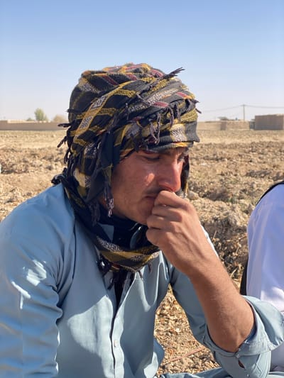 An outdoor, eye-level, daylight scene in Mandisar, Afghanistan, depicts a man, possibly of Afghan ethnicity, in a contemplative state. He is positioned in the foreground, facing partially to the right of the frame and looking downward. The man wears a light blue long-sleeved shirt, partially unbuttoned, and a dark, multi-colored headwrap or turban with patterns of black, yellow, and red, featuring fringed edges. His right hand is raised to his lower face, with fingers touching his chin/mouth area. The man's skin appears tanned, and he has short dark hair visible beneath the headwrap. Strong sunlight illuminates the right side of his face. To the immediate right, a portion of another individual wearing a white shirt is visible. The background consists of a dry, barren landscape with light-colored soil and sparse vegetation. In the far distance, under a clear blue sky, several low-rise structures, possibly mud-brick buildings, and a utility pole with wires are discernible on the horizon.