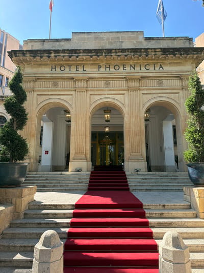 The image shows the exterior of the Hotel Phoenicia in Floriana, Malta. A wide, red carpet leads up a stone staircase to the hotel entrance. The entrance features a grand arched portico with classical columns. Above the portico, the words "HOTEL PHOENICIA" are inscribed in large, dark letters on a stone facade. Two flags, one with a red and white pattern and another with a blue and white design, are visible flying from poles on the roof against a clear blue sky. Two manicured, conical evergreen trees in large dark pots flank the staircase on either side. The architectural style is grand and imposing, with sandstone-colored stone prevalent in the building's construction. The scene is well-lit by sunlight, casting shadows on the stairs and the building. No people are visible in the image.