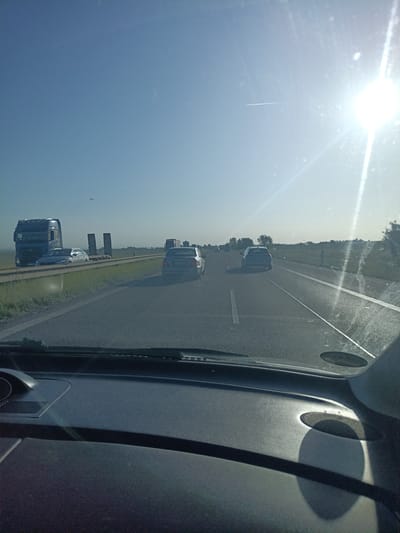 The image, taken from the perspective of a driver inside a vehicle, depicts a clear, sunny day on a highway. The highway appears to be in a rural or suburban setting, with grassy verges and a flat landscape stretching to the horizon. Several vehicles are visible, including a large dark truck on the left side of the road, a white car, and other cars further down the highway, suggesting moderate traffic. The sun is prominently positioned in the upper right quadrant of the image, casting a bright glare and creating lens flare effects on the windshield. A faint contrail from an airplane is visible in the sky. The foreground shows the interior of the vehicle, specifically the dashboard and windshield wipers. The location is identified as Svémyslice, Czechia.