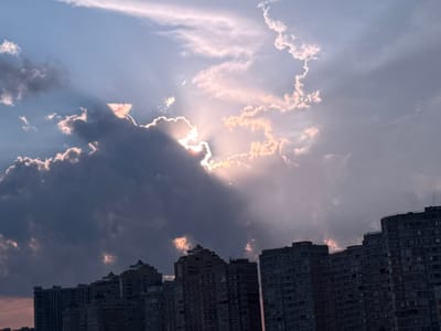 The image depicts a dramatic sunset over a cityscape in Starobzhegokay, Russia. The sky is filled with a mix of dark, ominous clouds and bright, illuminated patches where the sun is breaking through. Rays of sunlight emanate from behind a large cloud formation, casting a warm, ethereal glow and creating crepuscular rays that stretch across the sky. The sun itself is largely obscured, but its brilliance is evident from the light filtering through the clouds.

Silhouetted against the vibrant sky are the upper floors of several large, apartment buildings. These structures appear to be quite tall and are densely packed, suggesting an urban or suburban environment. The architecture seems typical of Soviet-era or modern Russian construction, with repetitive window patterns and flat roofs. The buildings are dark and lack individual detail due to the strong backlight from the sunset.

The time of day is clearly evening, as indicated by the sunset. The weather appears to be dynamic, with both dense cloud cover and clear skies coexisting, creating a visually striking contrast. The scene evokes a sense of awe and perhaps a touch of melancholy due to the dramatic lighting and the imposing, dark buildings. The location is confirmed as Starobzhegokay, Russia. There is no visible text in the image.