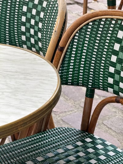 This is a close-up, eye-level, outdoor shot of two green and white woven bistro chairs and a portion of a round table in Bordeaux, France. The chairs have wooden frames and woven seats and backs. The table has a light-colored, possibly marble, top with a thin metal edge. The chairs are positioned around the table, with one chair in the foreground and another partially visible behind it. The background shows a cobbled stone ground. There are no people in the image.