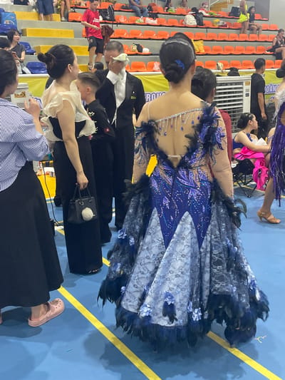 The image depicts a scene within the Binh Duong Sports Center, located in Thuận An City, Vietnam. The foreground is dominated by a woman in an elaborate, sparkly blue and white ballroom dancing dress adorned with feathers. Her back is to the camera, and the intricate details of the dress are visible, including sequins, beading, and floral appliqués. To the left of the woman in the dancing dress, another woman is visible from the side, wearing a black dress and holding a small black handbag with a fluffy keychain. She is partially obscured by a woman in a blue and white striped top, also in the foreground.
In the middle ground, several individuals are present. A man in a black tuxedo is wiping his face with a white cloth. Beside him, a young boy in a black outfit, possibly a dance costume, stands. Further back, other individuals are seated in orange tiered seating. One person is wearing a red t-shirt and dark shorts. In the background, a large white air conditioning unit is visible against a yellow banner. The floor is blue with distinct yellow lines. The overall impression is that of a competition or event related to ballroom dancing.