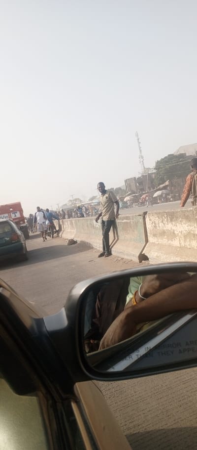 This image captures a street scene in Iddah, Nigeria, from the perspective of a vehicle's passenger side. The foreground is dominated by the vehicle's side mirror, reflecting part of the occupant's arms wearing a light green sleeve and wristbands, along with a blurred reflection of the vehicle's interior. A concrete road barrier runs diagonally from the lower left to the upper right, separating the viewer's immediate road from the opposite lane and a busy roadside area.
Standing prominently on the far side of this barrier, slightly right of center, is an adult male with dark skin, wearing a light-colored striped polo shirt and dark pants with sandals. He is facing generally towards the camera, with a direct gaze. His shadow is cast long on the barrier.
Beyond the barrier, a multi-lane road is visible with traffic. A red truck with "T" and "56 KM" markings is partially visible on the left. Several individuals are walking or operating motorcycles. Further back, a dense collection of people and structures suggests a market or commercial area. Multiple street vendors' umbrellas are visible among the crowd. A tall, white cellular communication tower stands in the distant background, against a hazy, bright sky. Other buildings, some under construction or in disrepair, are also visible in the background. The overall environment appears active and urban.