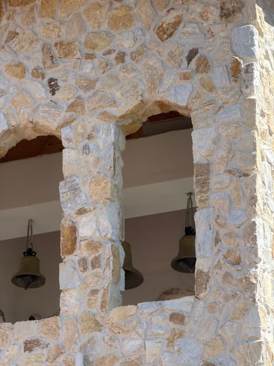 A close-up, ground-level exterior shot captures a section of a stone-built structure with two arched openings. The stonework is rustic, featuring irregularly shaped, rough-hewn stones in varying shades of beige, tan, and white, mortared together. One opening reveals a glimpse of a wooden roof structure above, while the other is an open archway.
Suspended from chains within the right-hand opening are two large, brass bells. The bell on the left is more clearly visible, showing a dark, ornate crown-like fixture at the top and a dark clapper hanging inside. The bell on the right is partially obscured. Below the bells, a segment of a light-colored ceiling is visible. Further down, near the bottom of the frame and peeking from behind the stonework, is a black, dome-shaped object that appears to be a security camera. The overall impression is that of a bell tower or part of a church or other significant building.
The image was taken outdoors, likely on a sunny day, given the bright lighting and shadows. The setting is the vicinity of the Church of St. Nicholas the Wonderworker in the town of Kocherinovo, Bulgaria.
