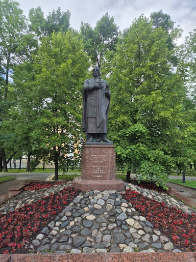 This is a vertical, eye-level, outdoor, medium shot of a bronze statue of Alexander Yaroslavich Nevsky on a granite pedestal. The statue depicts him in traditional clothing, holding a scroll in his left hand. The pedestal is inscribed with Russian text, which reads: "To Saint Alexander Yaroslavich Nevsky, from grateful descendants. 2010, the year of the 90th anniversary of the Republic of Karelia." The statue is situated in a park-like setting, with lush green trees and a flower bed filled with small red flowers and large stones. A black metal fence and a paved walkway are visible in the background. The lighting suggests it is daytime, possibly overcast. The overall scene evokes a sense of remembrance and historical appreciation.
