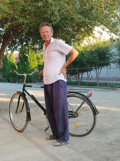 An elderly man stands next to a dark green bicycle in an outdoor setting. He is wearing a white short-sleeved button-up shirt and dark blue loose-fitting pants. He has his left hand on his hip and his right hand is holding the handlebars of the bicycle. The bicycle has a silver handlebar and a silver bell. The man is wearing dark gray sandals. The ground is a light-colored, textured surface, possibly concrete or dirt, with some shadows cast by a large tree with green leaves overhead. In the background, there are green bushes and trees, a light-colored fence, and a building with a red roof. The scene appears to be in a residential area. The location of Shovot, Uzbekistan, is provided as context for the image.