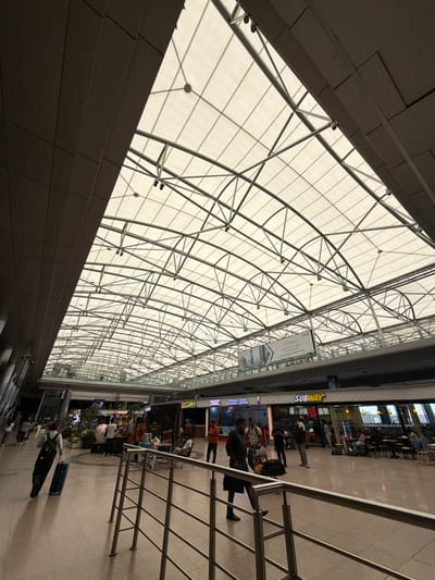 The image shows an interior view of Rajiv Gandhi International Airport in Hyderabad, India. The expansive ceiling is constructed with a translucent white fabric supported by a complex steel framework, allowing diffused natural light to illuminate the space. The airport terminal features a polished, light-colored floor with visible expansion joints. In the foreground, a person is pulling a rolling suitcase. The middle ground shows a row of shops, including a Subway restaurant, with seating areas and patrons. Other people are visible walking through the terminal, some with luggage. The environment appears to be a busy, well-lit airport concourse.
