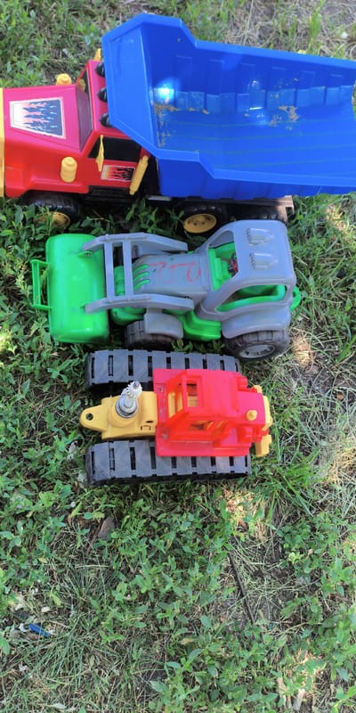 Three toy construction vehicles are arranged on a grassy area in Esipovo, Russia. The largest toy, a red and blue dump truck, is positioned at the top of the image. Below it is a green and gray toy front-loader, and at the bottom is a red and yellow toy bulldozer with tracks. The vehicles are likely outdoors, with the grass and dappled sunlight indicating a natural environment. There are no people or animals visible in the scene. The focus is solely on the toys.
